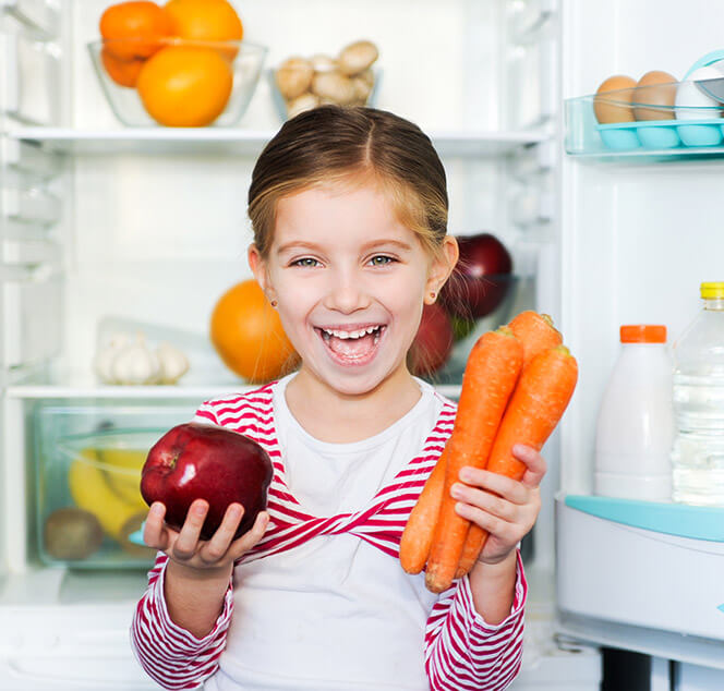 女孩站在打開的冰箱前，手裡拿著紅蘿蔔和蘋果開心笑著